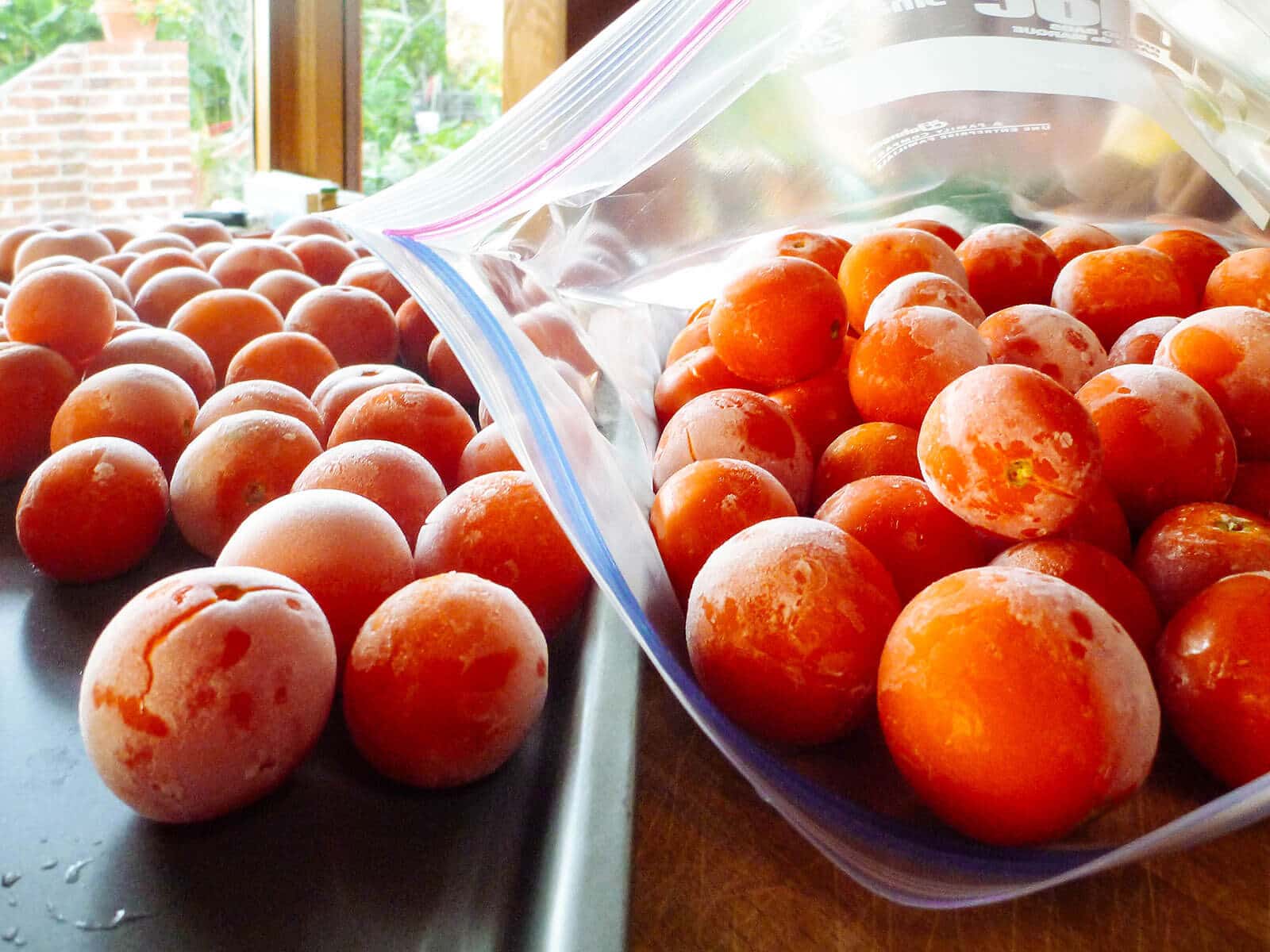 Baking sheet of frozen whole tomatoes next to Ziploc bag of frozen fresh tomatoes