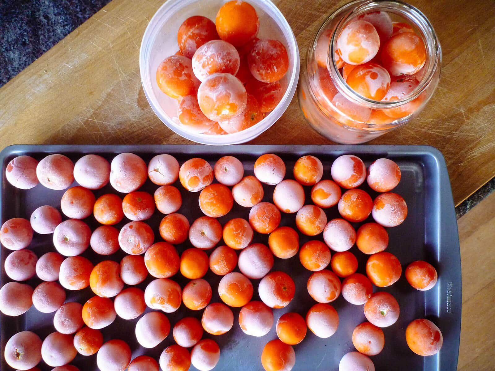 Frozen whole cherry tomatoes on a sheet pan, next to two jars filled with more frozen cherry tomatoes on a kitchen counter