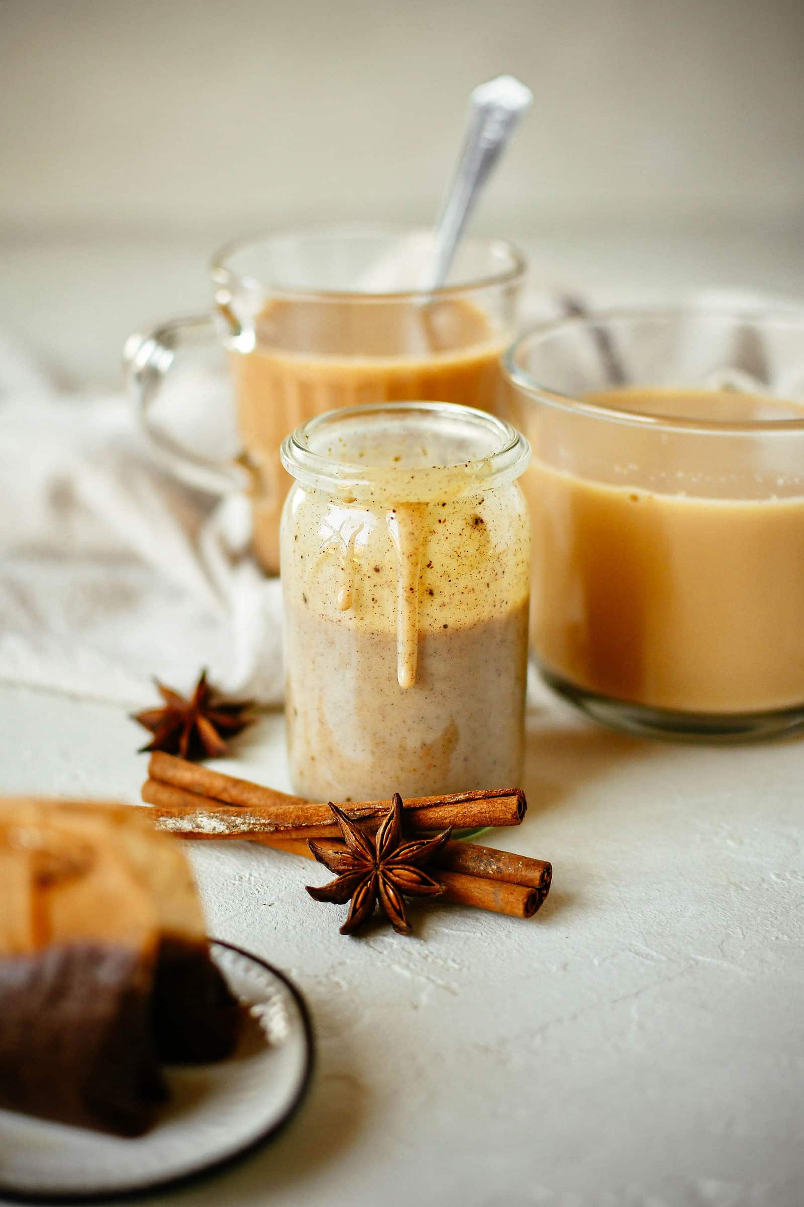 A jar of homemade chai concentrate sitting in front of two mugs of chai, with cinnamon sticks, star anise, and brewed tea bags in the foreground